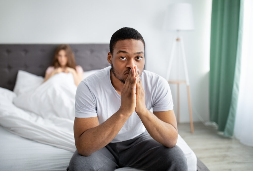 Young guy feeling unhappy, sitting on bed deep in thought, his disappointed wife on background. Multiracial family having fight, thinking about divorce or separation. Negative emotions, problems